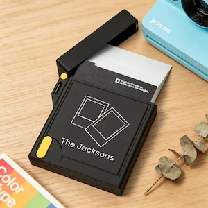 May include: A black square film storage case with a hinged lid, featuring a white graphic of two overlapping instant photos and the text The Jacksons. The case has a small yellow accent button and contains a stack of white instant film sheets.