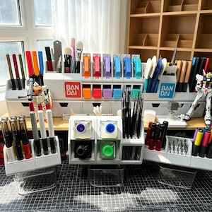 May include: A modular grey plastic desk organiser system featuring multiple compartments for art supplies, paint brushes, and markers. The units display the Bandai logo and include small drawers, clear storage bins, and various hobby tools.