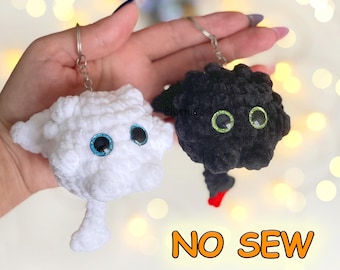 Può includere: Due portachiavi in peluche realizzati in filato morbido, uno bianco con occhi blu e uno nero con occhi verdi e una piccola punta della coda rossa. Il portachiavi nero ha un'etichetta con scritto PlushCo. Il testo NO SEW è stampato in arancione.