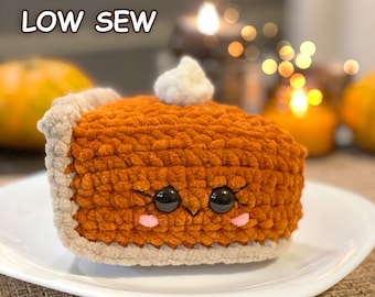 Può includere: Un peluche all'uncinetto a forma di fetta di torta di zucca con crosta beige, ripieno arancione e un ciuffo di panna montata bianca sopra. La torta ha due grandi occhi neri di sicurezza, ciglia ricamate e guance rosa. Testo LOW SEW.