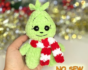 Può includere: Una bambola di peluche verde chiaro realizzata in filato morbido con una finitura testurizzata. La bambola presenta occhi in plastica nera, sopracciglia ricamate e un piccolo sorriso. Indossa una sciarpa a righe rosse e bianche. Testo NO SEW.