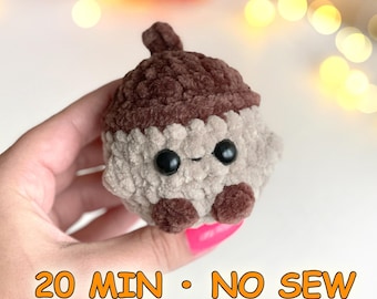 Può includere: Un piccolo giocattolo di peluche a forma di ghianda lavorato all'uncinetto con un cappuccio marrone scuro e un corpo beige chiaro. Presenta due occhi di sicurezza in plastica nera e due piccoli piedi marrone scuro. Il testo 20 MIN • NO SEW è in basso.
