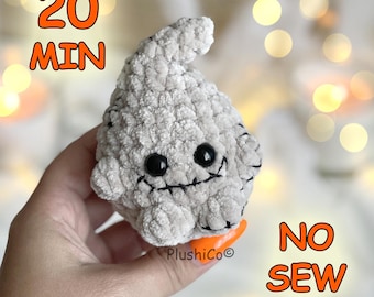 Può includere: Un piccolo fantasma di peluche grigio chiaro con una consistenza morbida e soffice, occhi di sicurezza in plastica nera e una bocca nera cucita. Il testo 20 MIN e NO SEW è visibile in arancione, insieme al testo PlushiCo in basso.