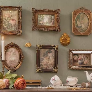 May include: A collection of eight ornate gold and dark wood picture frames containing floral and landscape art, arranged on a wall above a table featuring a white teapot, teacups, a lit candle in a metal holder, and several artificial roses.
