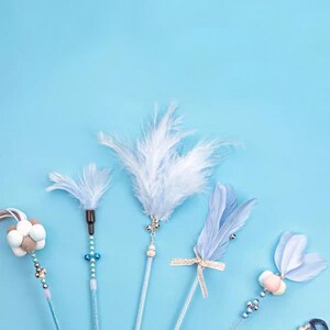 May include: A selection of cat toys with feather and ribbon details. The toys feature light blue and white feathers, decorative beads, and ribbons. The toys are set against a light blue backdrop.