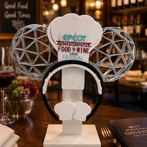 May include: A headband featuring silver geometric ears and a white chef hat centerpiece with the text EPCOT INTERNATIONAL FOOD & WINE 2020 FESTIVAL, accompanied by a pair of matching white chef hat earrings displayed on a white stand.
