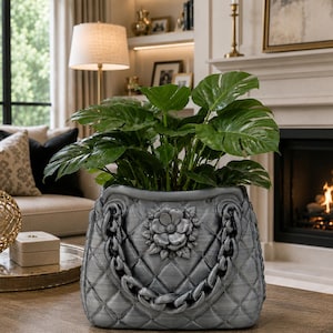 May include: A gray planter shaped like a handbag with a quilted diamond pattern, a central sculpted flower emblem, and a decorative chain link detail draped across the front. The planter holds a lush green plant with large, split leaves.