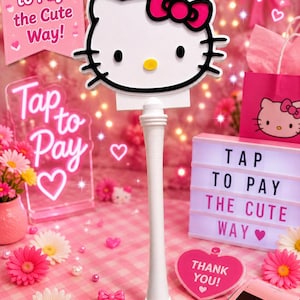 May include: A white Hello Kitty sign with a pink bow, black eyes, and yellow nose mounted on a white cylindrical stand. The sign features the text Tap to Pay... the Cute Way! and is surrounded by pink decor, a light box, and a payment terminal.