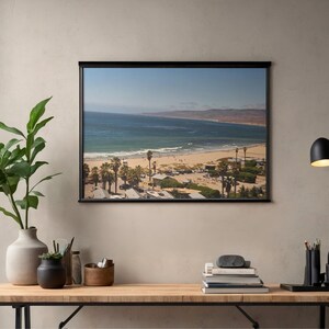 May include: A rectangular wall art print featuring a coastal landscape with a sandy beach, blue ocean, and distant hills. The scene includes palm trees, parked vehicles, and small structures near the shoreline, all framed in a thin black border.