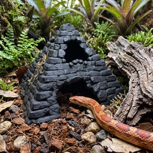 May include: A black, pyramid-shaped reptile hide with a textured, brick-like exterior and two arched openings. The structure features a tiered design with a wide base and a pointed top, providing a sheltered space for small animals to rest inside.