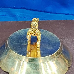 Puede incluir: Una pequeña figura de metal pulido color oro de una deidad en posición de gateo, montada en el centro de un plato circular de metal con borde festoneado. La figura presenta rasgos faciales detallados y un pequeño peinado de moño.