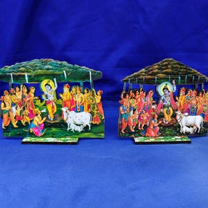 Puede incluir: Dos recortes decorativos de madera que representan la escena de Govardhan Puja. Cada pieza presenta una figura central de piel azul levantando una montaña, rodeada por una multitud con vestimenta tradicional colorida, una vaca blanca y un ternero.