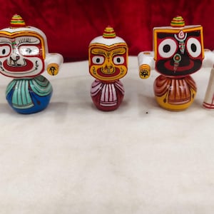 May include: Three hand-painted wooden figurines featuring stylised faces with large circular eyes. The left figure has a blue base, the centre figure is smaller with a pink base, and the right figure has a yellow base and a rectangular head shape.