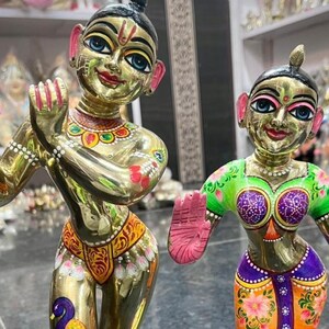 May include: Two gold-coloured statues of deities, each standing on a lotus flower base. The statues are adorned with colourful painted details, including peacock feather designs and floral patterns. One statue has hands in a prayer position.