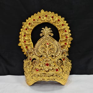 May include: A gold-colored crown featuring an ornate, embossed floral and leaf pattern. The headpiece is accented with small, circular red gemstones embedded throughout the design, including a central flower motif and a circular halo frame.