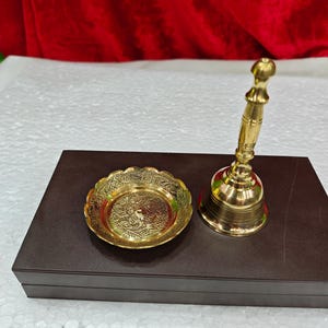 May include: A polished brass hand bell with a slender, tiered handle and a matching circular brass dish featuring an intricate, embossed floral pattern along the scalloped rim, both resting on a dark brown rectangular box.