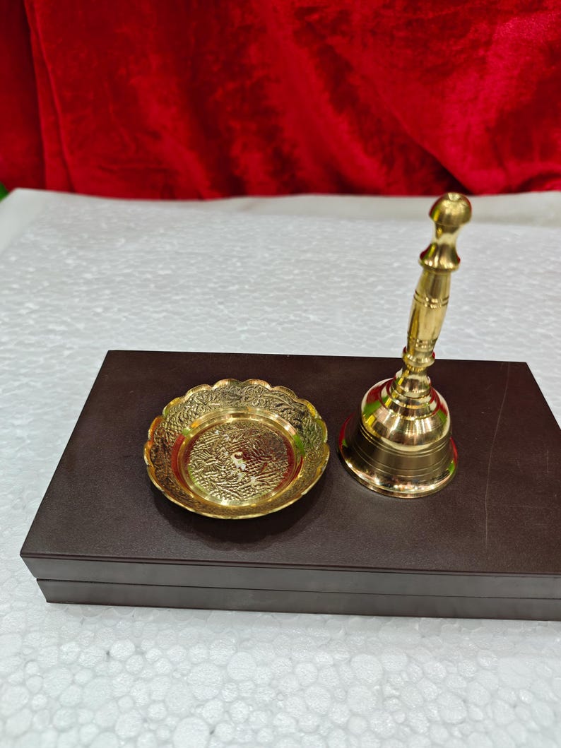 May include: A polished brass hand bell with a slender, tiered handle and a matching circular brass dish featuring an intricate, embossed floral pattern along the scalloped rim, both resting on a dark brown rectangular box.
