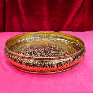 May include: A round, polished gold-coloured metal tray featuring an intricate, embossed geometric pattern on the flat interior surface and a decorative, repeating scalloped design along the raised outer rim, creating a textured, reflective finish.