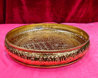 May include: A round, polished gold-colored metal tray featuring an intricate, embossed geometric pattern on the flat interior surface and a decorative, repeating scalloped design along the raised outer rim, creating a textured, reflective finish.
