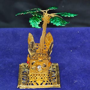 May include: A gold-colored metal decorative object featuring a square base with intricate filigree patterns, a central box-like structure adorned with a flower and a clear stone, and a palm tree top with metallic green leaves and a textured trunk.