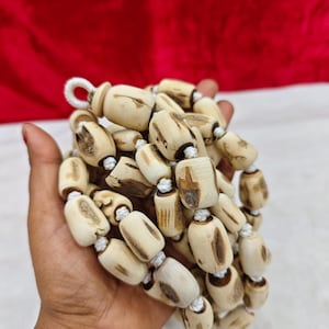 May include: A strand of cylindrical beads made from light-colored wood with natural dark brown grain patterns and knots. The beads are strung together on a white cord, creating a textured, organic appearance with varied shapes and sizes.