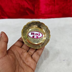 May include: A small, round, gold-colored brass dish with a scalloped edge and intricate embossed floral patterns. A white oval sticker with a purple border is centered on the dish, featuring the text "Raj 2" in bold, dark purple lettering.