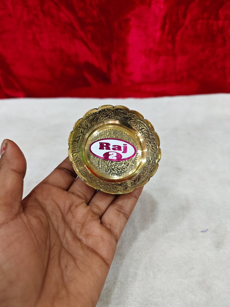 May include: A small, round, gold-colored brass dish with a scalloped edge and intricate embossed floral patterns. A white oval sticker with a purple border is centered on the dish, featuring the text "Raj 2" in bold, dark purple lettering.