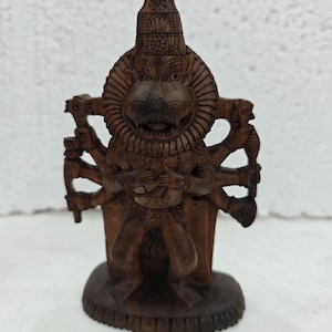 May include: A dark brown wooden sculpture of a multi-armed deity with a lion head, featuring intricate carvings on the face, mane, and tall headdress. The figure stands on an oval base, with multiple arms extended, holding various objects.