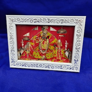 Puede incluir: Un marco blanco rectangular con un patrón floral en relieve que contiene una impresión vibrante de dos deidades. La figura central toca una flauta, vestida con prendas amarillas y rojas brillantes, flanqueada por una figura más pequeña.
