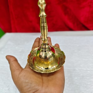 May include: A polished brass hand bell featuring a tall, slender handle and a wide, flared base with intricate embossed floral patterns along the rim. The bell has a smooth, reflective metallic finish and a classic, symmetrical design structure.