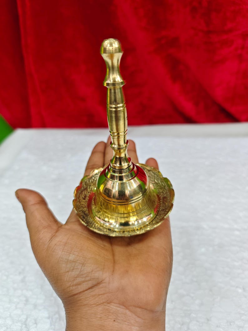 May include: A polished brass hand bell featuring a tall, slender handle and a wide, flared base with intricate embossed floral patterns along the rim. The bell has a smooth, reflective metallic finish and a classic, symmetrical design structure.