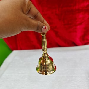May include: A polished brass hand bell featuring a slender, cylindrical handle with horizontal grooved detailing and a flared, smooth bell body. The metal surface has a reflective, metallic gold finish that catches the light, showcasing a classic design.