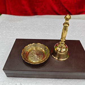 May include: A polished brass hand bell with a slender handle and a matching small circular brass dish featuring an intricate embossed floral pattern. Both items are metallic gold in color and display a reflective, smooth finish on their surfaces.