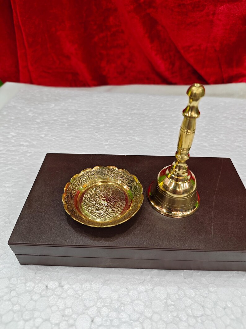 May include: A polished brass hand bell with a slender handle and a matching small circular brass dish featuring an intricate embossed floral pattern. Both items are metallic gold in color and display a reflective, smooth finish on their surfaces.
