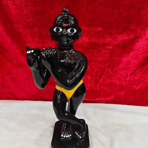 May include: A glossy black figurine of a standing figure with large white eyes, a small red dot on the forehead, and a yellow loincloth. The figure is posed with arms raised as if playing a flute, standing on a rectangular black base.