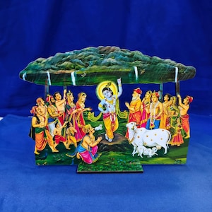 May include: A decorative wooden cutout depicting the Hindu deity Krishna lifting Govardhan Hill. The scene features Krishna in blue skin with yellow garments, surrounded by villagers, a white cow, and a calf, all sheltered under the mountain.