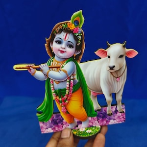 Op de afbeelding: Een platte, uitgesneden decoratieve figuur met een jonge godheid met een blauwe huid die een houten fluit bespeelt, versierd met een pauwenveer en bloemenslingers, naast een witte koe op een voetstuk met paarse en groene bloemmotieven.