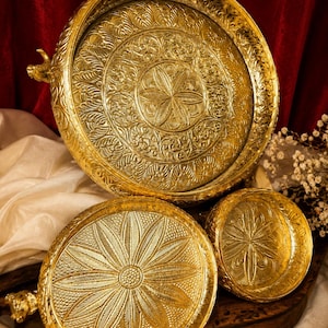 Pode incluir: Um conjunto de três tabuleiros redondos de metal dourado com padrões intrincados de flores e folhas em relevo. O tabuleiro maior tem um design floral central, enquanto os dois menores exibem pétalas detalhadas e pegas em forma de animal.