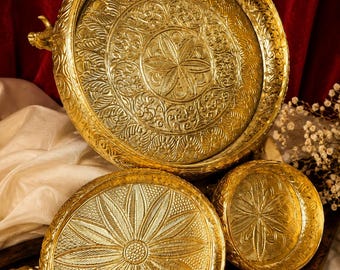 May include: A set of three round gold-colored metal trays featuring intricate embossed floral and leaf patterns. The largest tray has a prominent central flower design, while the two smaller trays display detailed petal motifs and small animal-shaped handles.