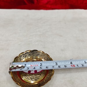 May include: A round brass dish with an intricate embossed floral pattern along the scalloped rim. A measuring tape placed across the center shows a diameter of approximately 3.5 inches. The dish has a polished gold-tone finish.