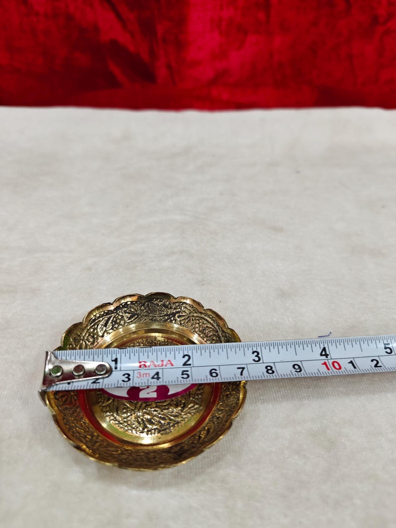 May include: A round brass dish with an intricate embossed floral pattern along the scalloped rim. A measuring tape placed across the center shows a diameter of approximately 3.5 inches. The dish has a polished gold-tone finish.