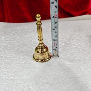 May include: A polished brass hand bell with a slender, tiered handle and a flared base. A vertical measuring tape placed next to the bell indicates a total height of approximately 4.5 inches from the base to the top of the handle.