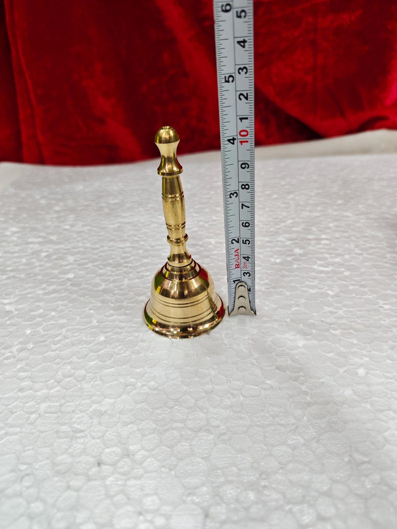 May include: A polished brass hand bell with a slender, tiered handle and a flared base. A vertical measuring tape placed next to the bell indicates a total height of approximately 4.5 inches from the base to the top of the handle.