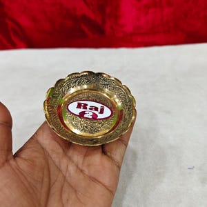 May include: A small, round, gold-colored brass bowl with a scalloped edge and intricate embossed floral patterns. The center of the bowl features a white oval sticker with a red border containing the text Raj 2 in bold red lettering.