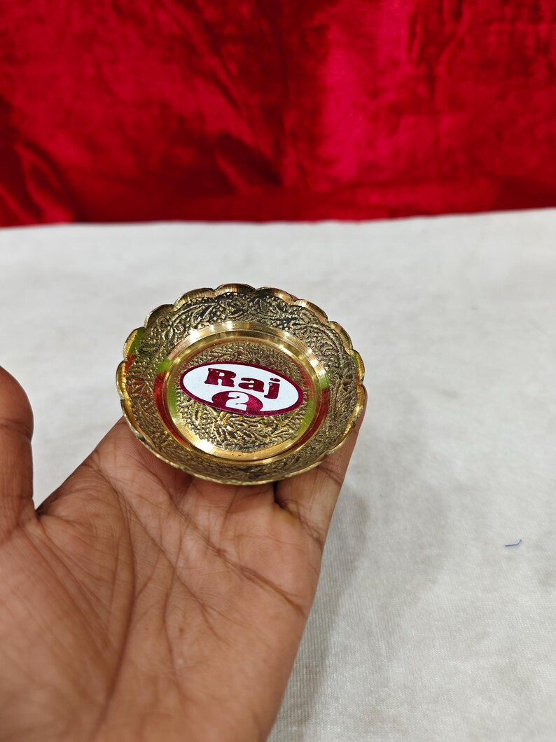May include: A small, round, gold-colored brass bowl with a scalloped edge and intricate embossed floral patterns. The center of the bowl features a white oval sticker with a red border containing the text Raj 2 in bold red lettering.