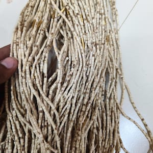 Puede incluir: Un conjunto de largas hebras de pequeñas cuentas cilíndricas de color blanco roto con patrones de veta de madera marrón, intercaladas con finos espaciadores tubulares de color oro metálico, dispuestas en un denso haz vertical.