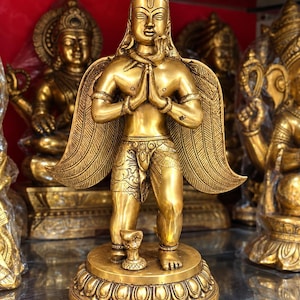 May include: A brass-coloured standing statue of a winged figure with hands pressed together in a prayer position. The figure features a snake coiled around its neck, detailed wing patterns, and stands on a circular base with a small snake figure.