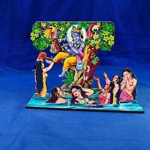 Puede incluir: Una decoración de madera recortada en varias capas que muestra a una figura de piel azul tocando la flauta sentada en un árbol, rodeada por varias figuras en el agua. La escena incluye follaje verde vibrante, prendas amarillas y tonos rosas.