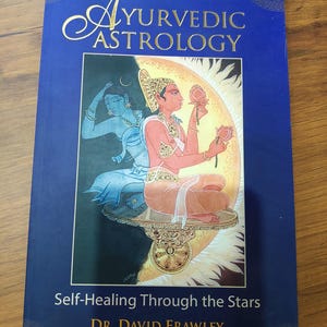 Könnte beinhalten: Ein blaues Buch mit dem Titel Ayurvedic Astrology: Self-Healing Through the Stars von Dr. David Frawley. Das Cover zeigt eine zentrale Illustration von zwei Figuren, eine blau und eine pfirsichfarben, die Lotusblumen vor Sonne und Mond halten.