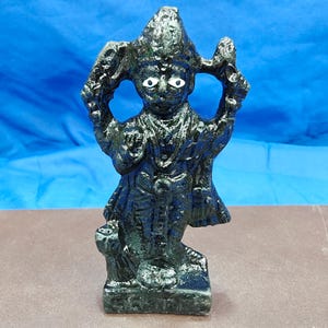 May include: A dark, textured, metallic-finish figurine of a standing deity with a pointed headdress, raised arms, and distinct white oval eyes. The figure is depicted in a frontal pose, wearing traditional attire with carved details on a base.